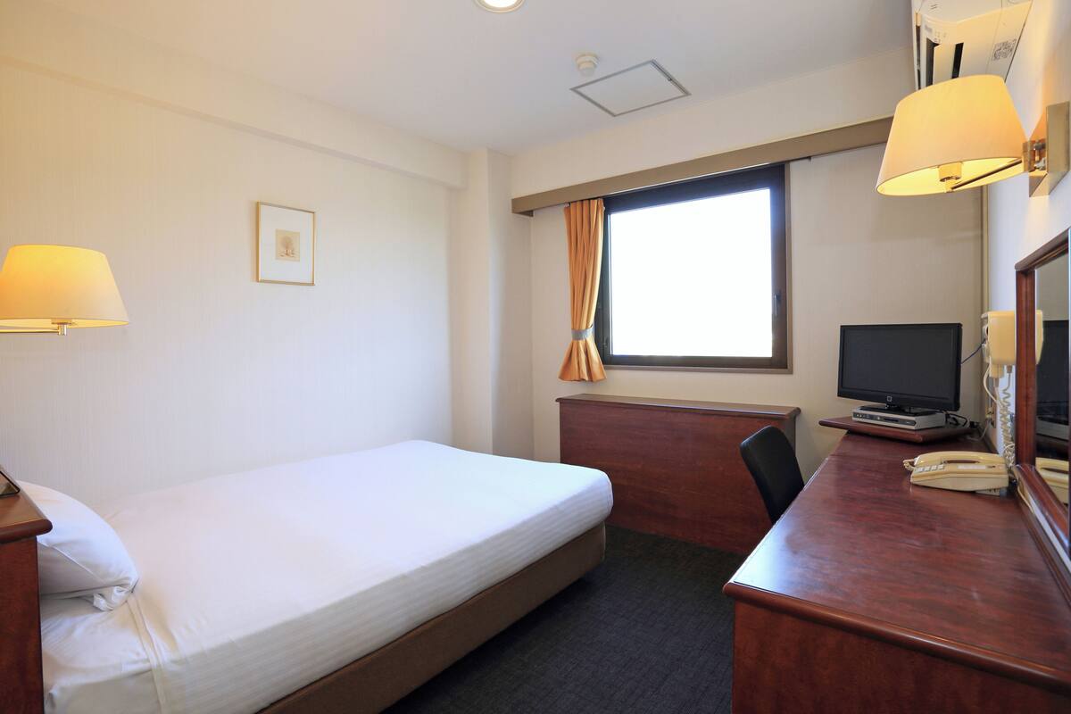 Habitació d'hotel amb llit, escriptori, cadira, televisió i finestra amb cortines.