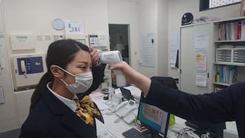 A person using a handheld infrared thermometer on another individual in an office setting with multiple phones, a computer, and various papers on the desk.