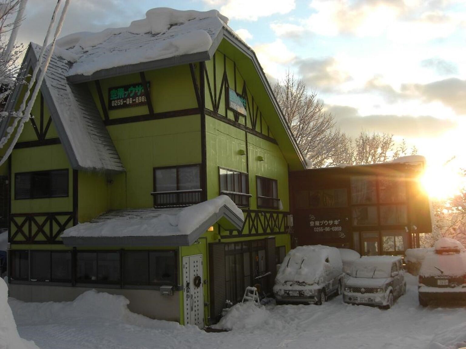 Un edificio de dos pisos con fachada verde y un balcón, rodeado de coches cubiertos de nieve.