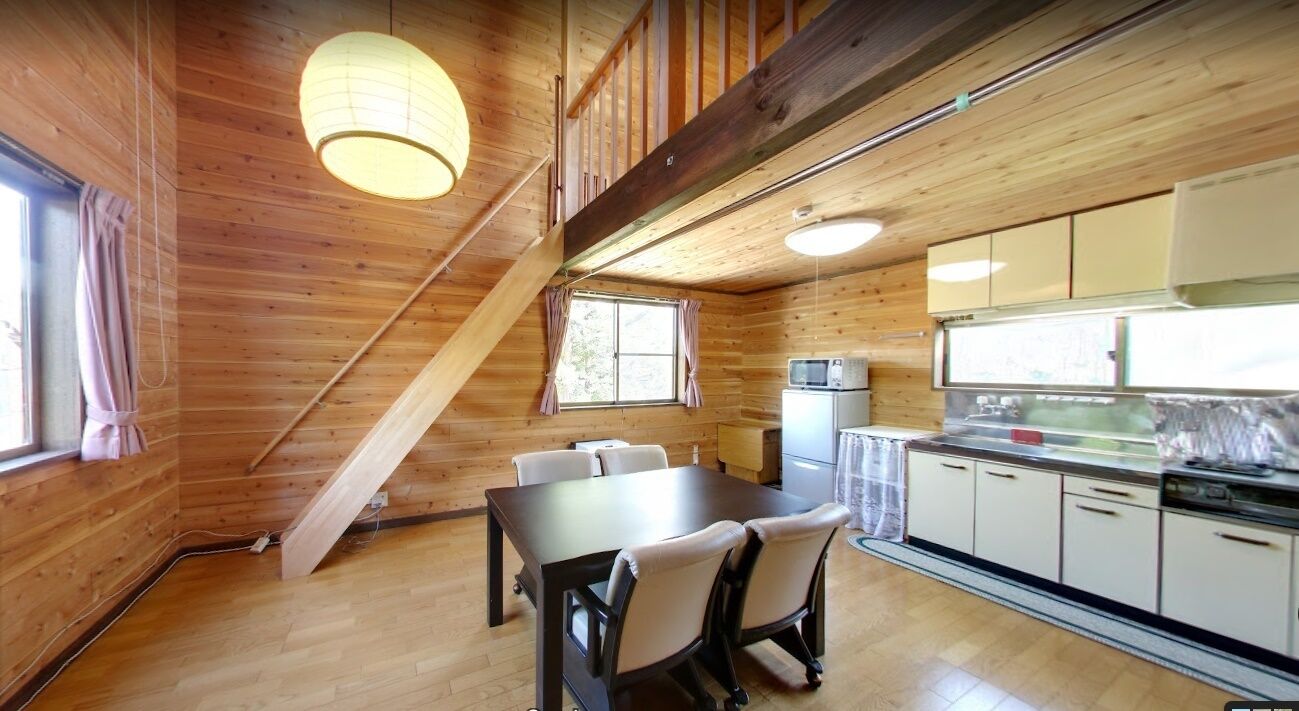 Interior de una cabaña de madera con una mesa de comedor, sillas, una cocina con armarios, un microondas y una ventana con cortinas.