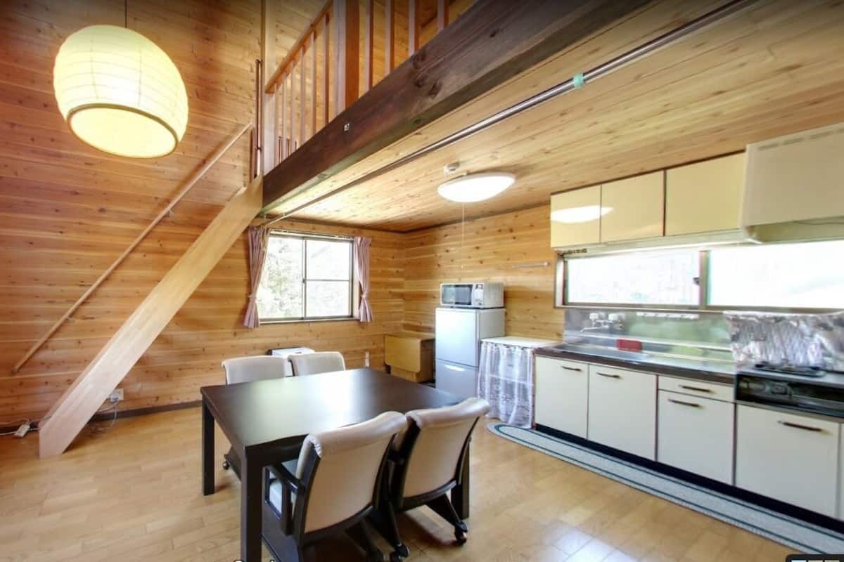 A wooden cabin interior with a dining table, chairs, a kitchen with cabinets, a microwave, and a window with curtains.