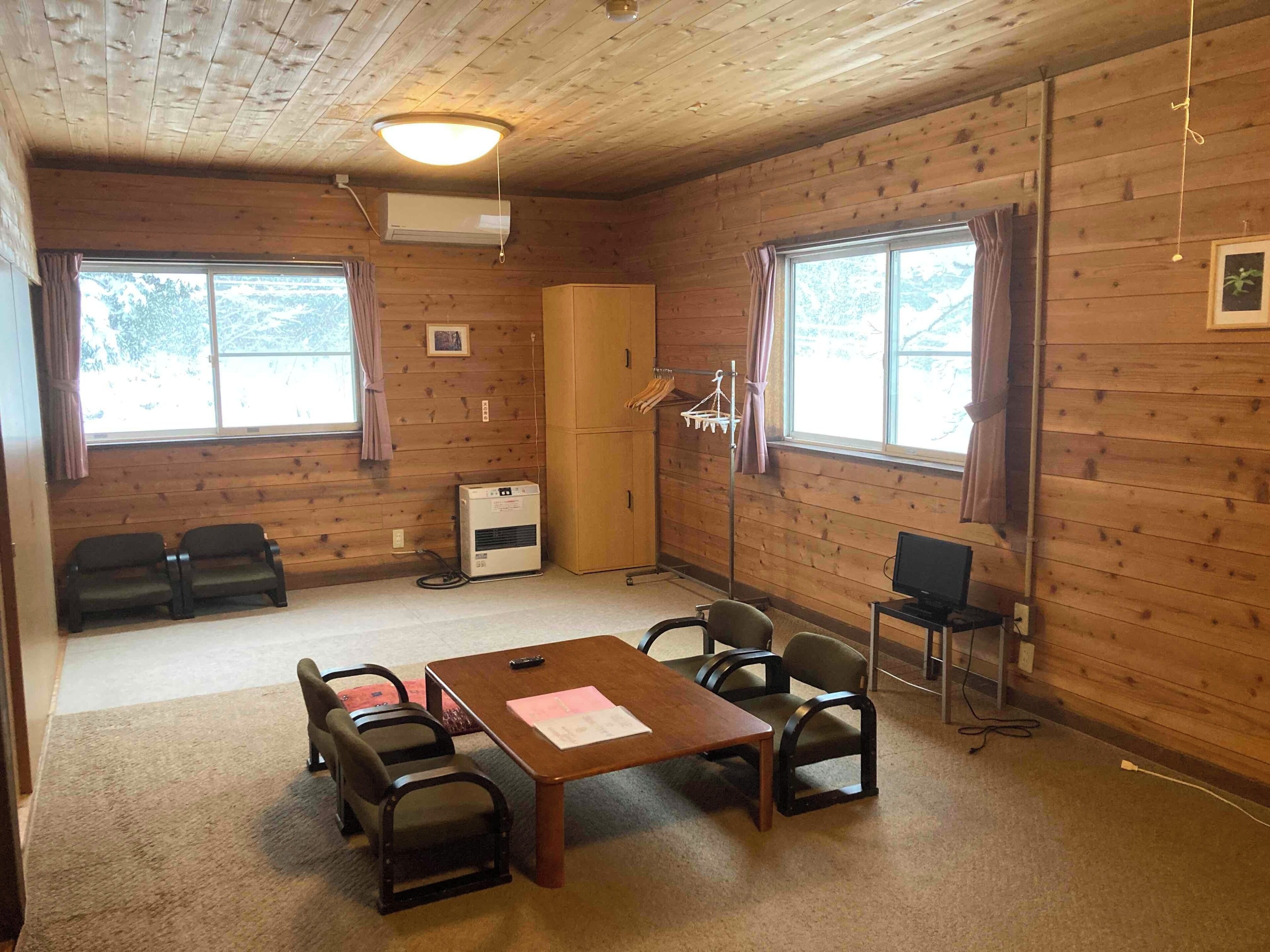 A room with wooden walls, a table with chairs, a television, an air conditioning unit, and windows with curtains.