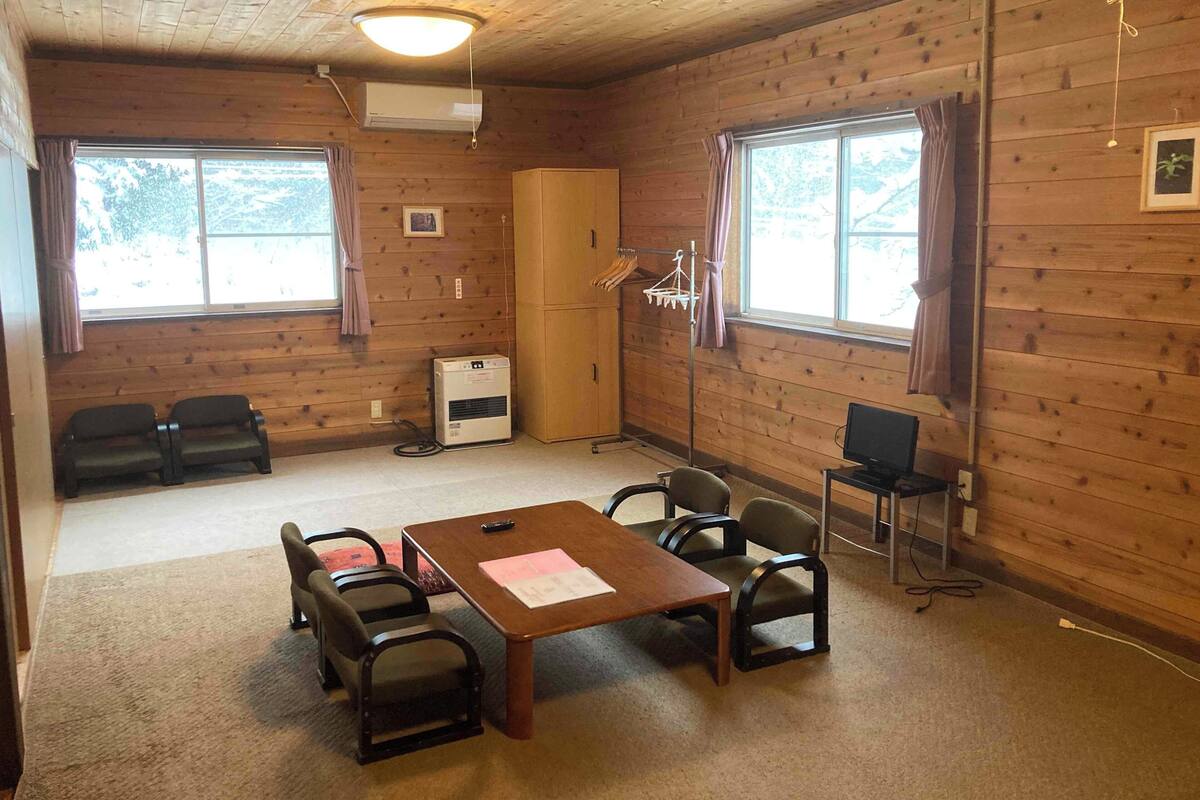 A room with wooden walls, a table with chairs, a television, an air conditioning unit, and windows with curtains.