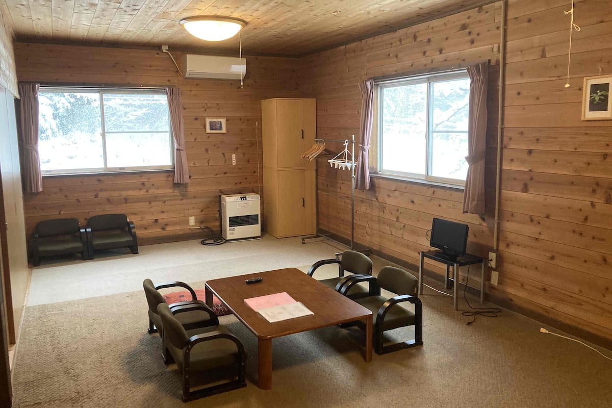 A room with wooden walls, a table with chairs, a TV, an air conditioning unit, and a window with curtains.