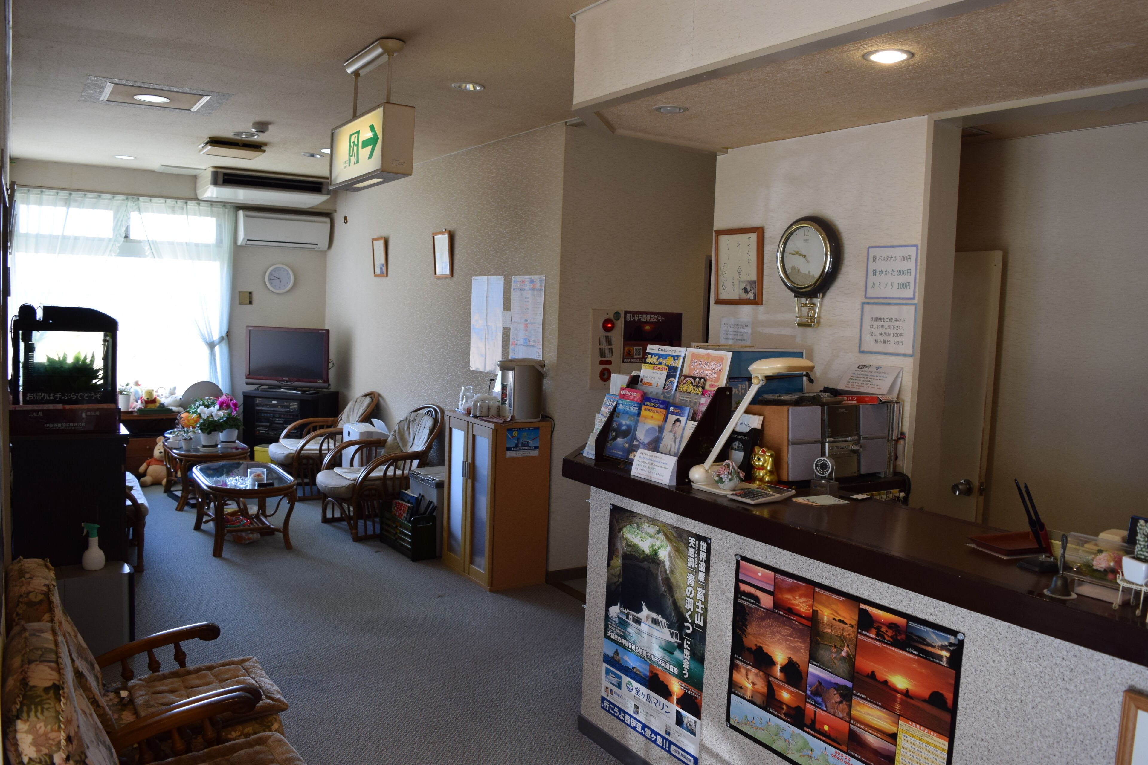 A hotel reception area with a counter displaying brochures and a clock. There are several chairs and a television in the background.
