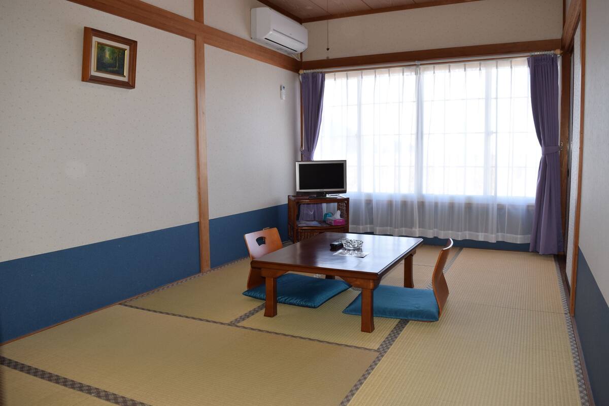 Een traditionele Japanse kamer met tatami vloerbedekking, een lage tafel met kussens, een televisie en een raam met gordijnen.