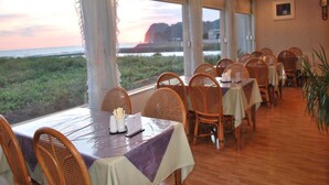 A restaurant with a view of the ocean at sunset, featuring wicker chairs, tables with tablecloths, and a wooden floor. - Seaside Resort Pension Surfrider (Nishiizu)