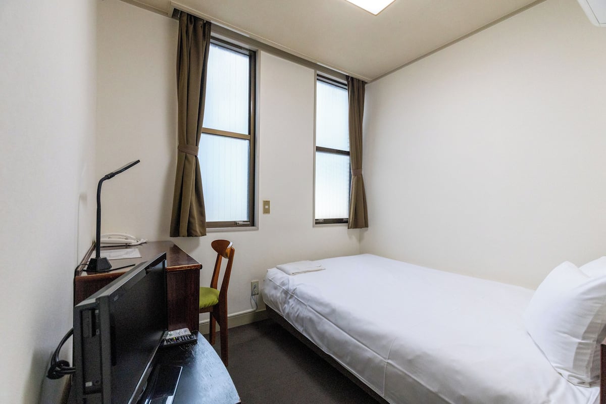 A small, clean room with a single bed, a desk with a computer, and two windows with curtains.