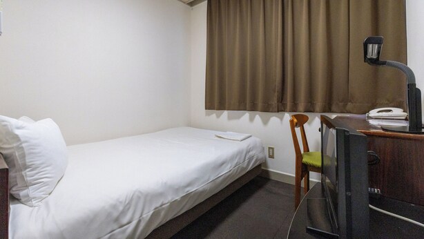 Uma cama de solteiro com roupa de cama branca, uma cadeira de madeira com almofada verde, uma mesa com telefone e abajur, e uma janela com cortinas castanhas.