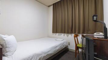 Un lit simple avec une literie blanche, une chaise en bois avec un coussin vert, un bureau avec un téléphone et une lampe, et une fenêtre avec des rideaux marron.