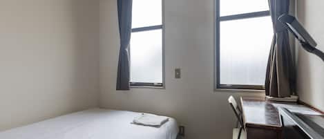 Une petite chambre avec un lit simple, un bureau et une chaise, deux fenêtres avec des rideaux et une prise de courant visible.