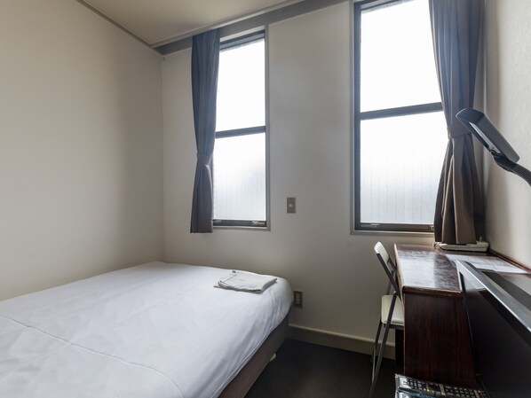 A small, single-bed room with a desk and chair, two windows with curtains, and a visible power outlet.