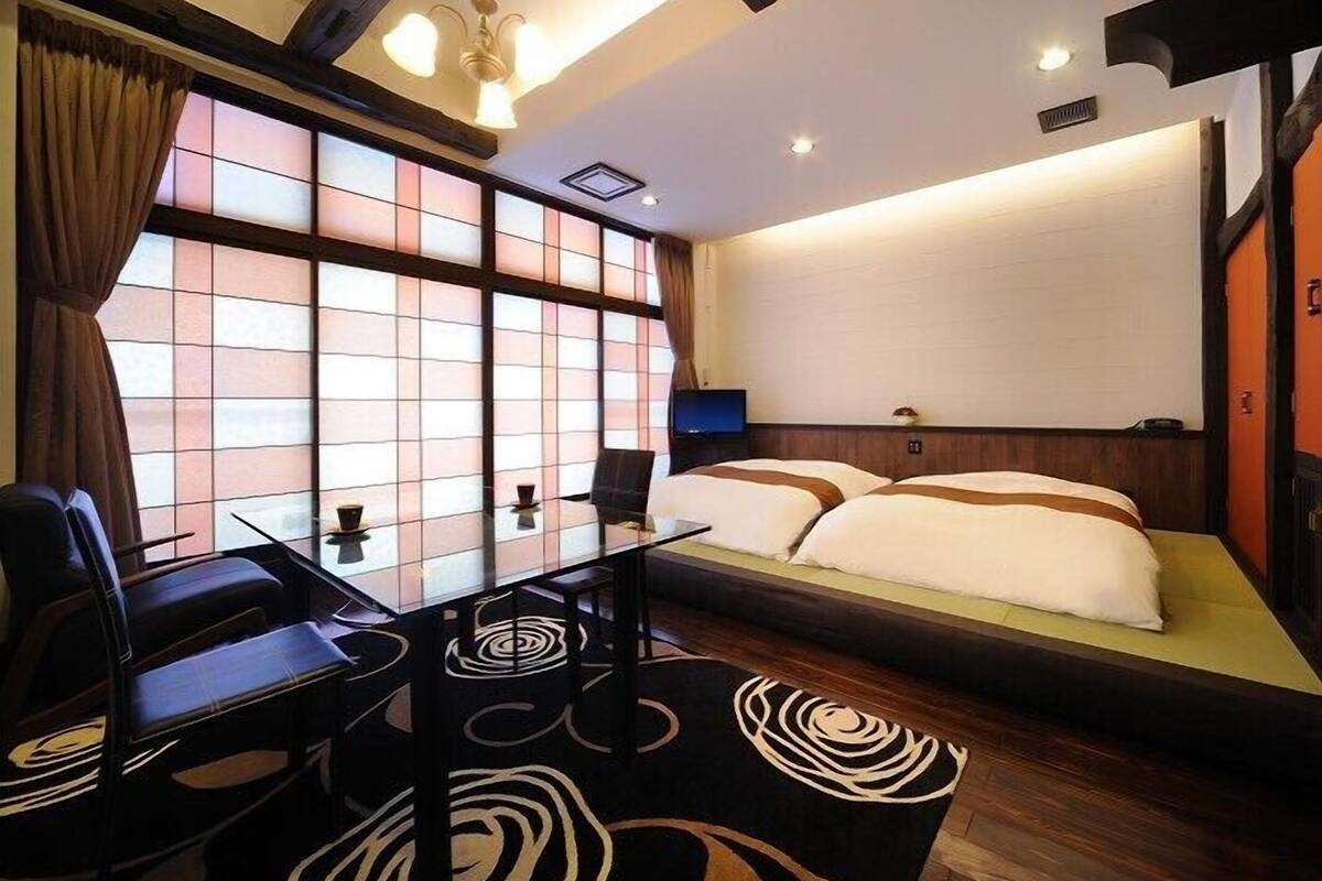 A hotel room with a traditional Japanese-style sliding door, a bed with a white and brown bedspread, a dining table with chairs, and a modern chandelier.