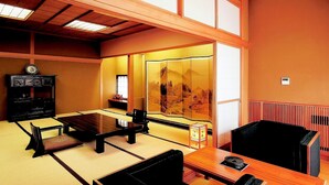 Una habitación tradicional japonesa con suelo de tatami, una mesa baja, una silla negra, un armario con un reloj y una puerta corredera con vistas a un paisaje montañoso.