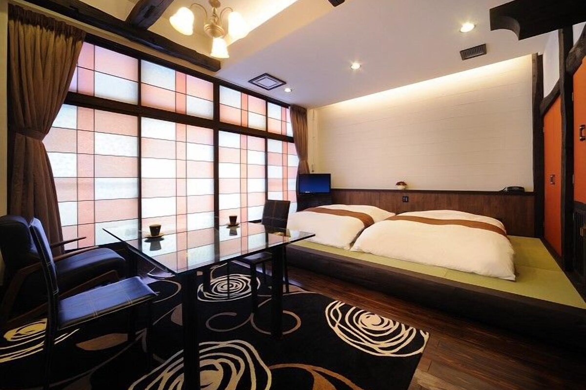 A hotel room with a traditional Japanese-style sliding door, a bed with a white and brown bedspread, a dining table with chairs, and a modern chandelier.