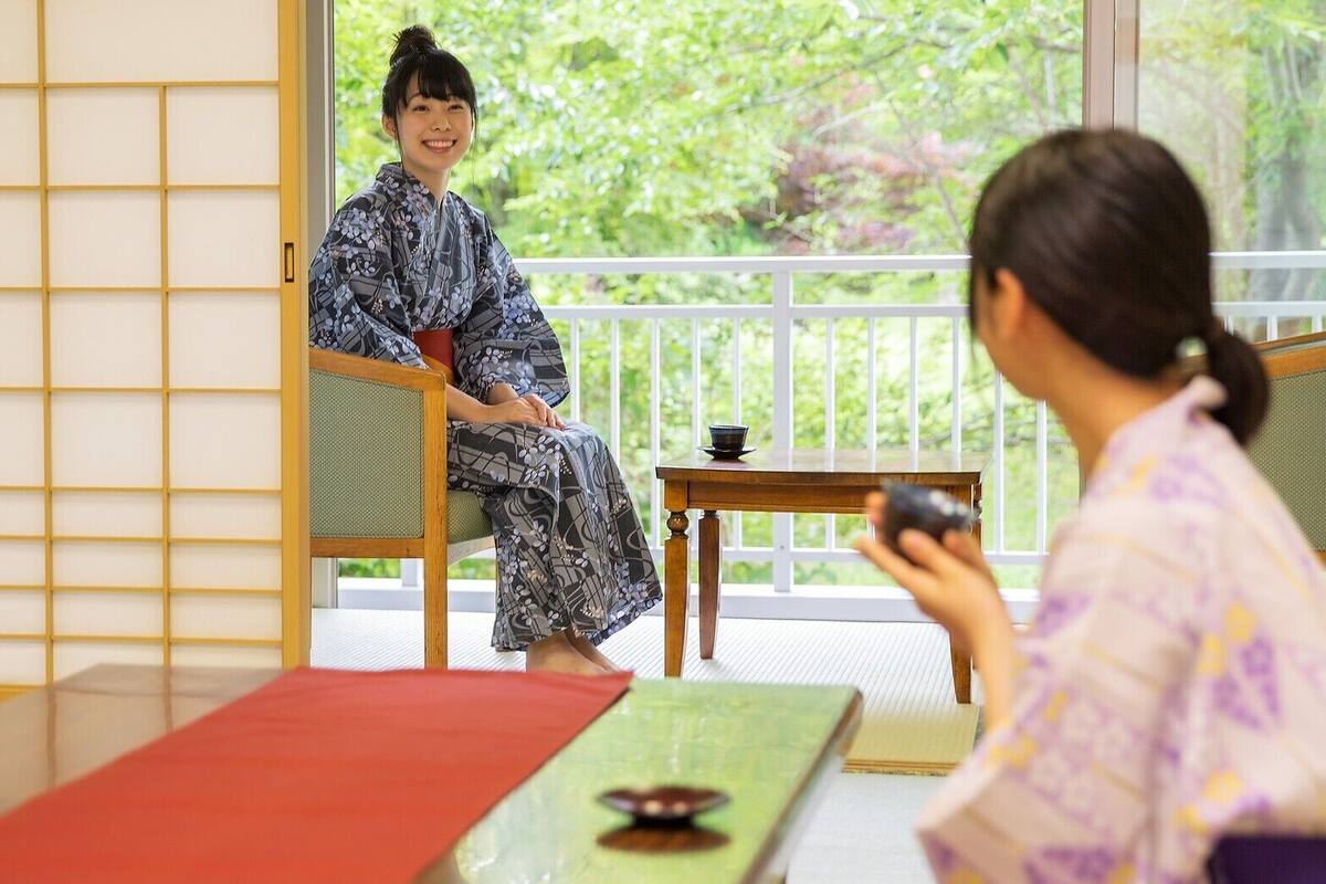 A traditional Japanese room with tatami flooring, a low table, and a sliding door leading to a balcony with a view of greenery.