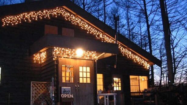 Una baita di legno con veranda, illuminata da luci a catena e un cartello che indica l'apertura.