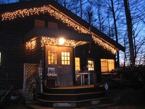 A wooden cabin with a porch, illuminated by string lights, and a sign indicating it is open. - Lodge Stack Point (Narusawa)