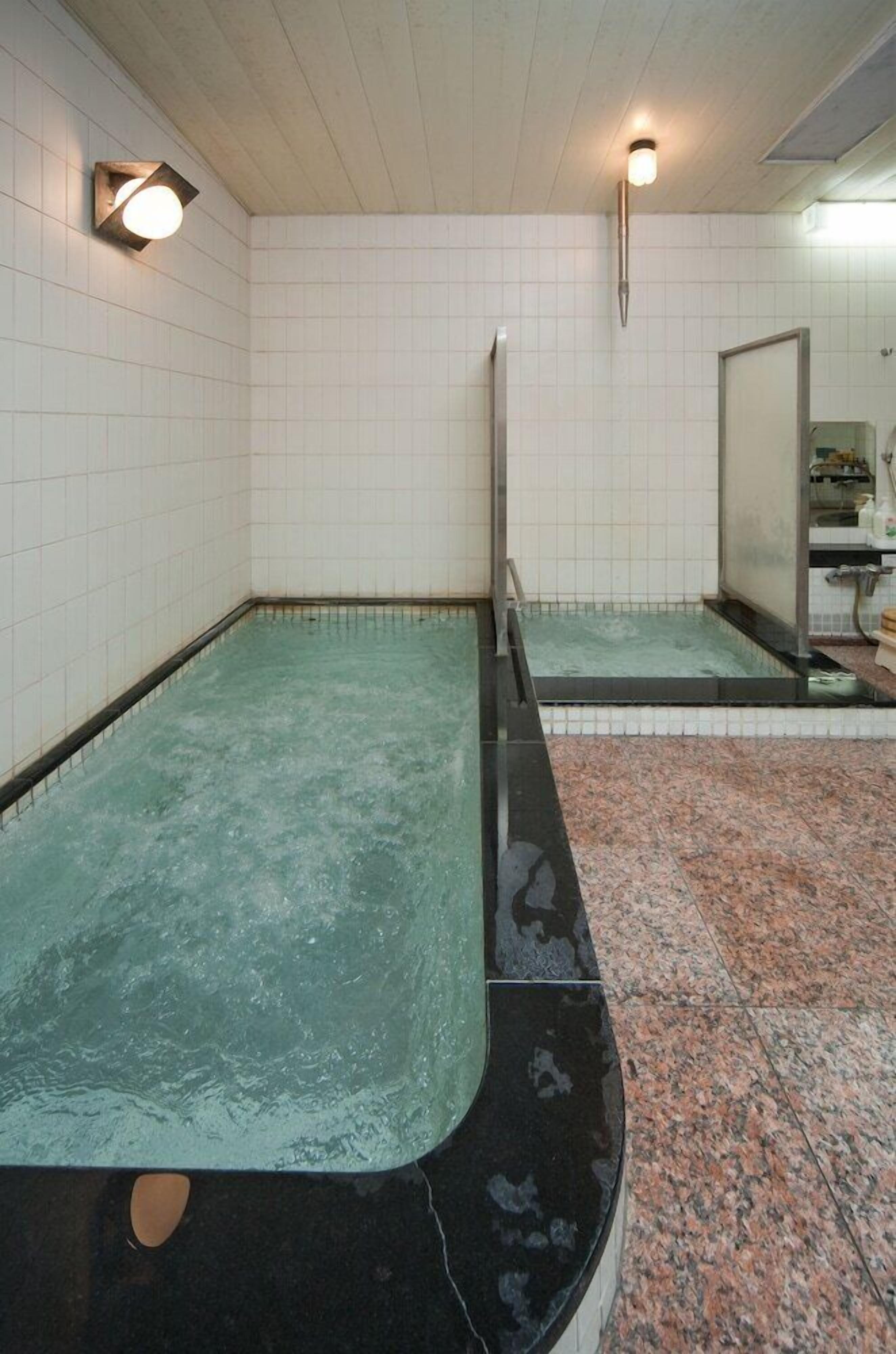 Ein Wellnessbereich mit einem rechteckigen Whirlpool, gefliesten Wänden und einem Spiegel, der den Raum reflektiert.