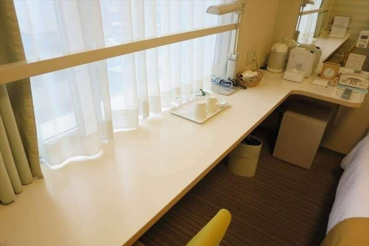 Une chambre d’hôtel comprenant un lit, un bureau avec une chaise, une fenêtre avec des rideaux et une poubelle.