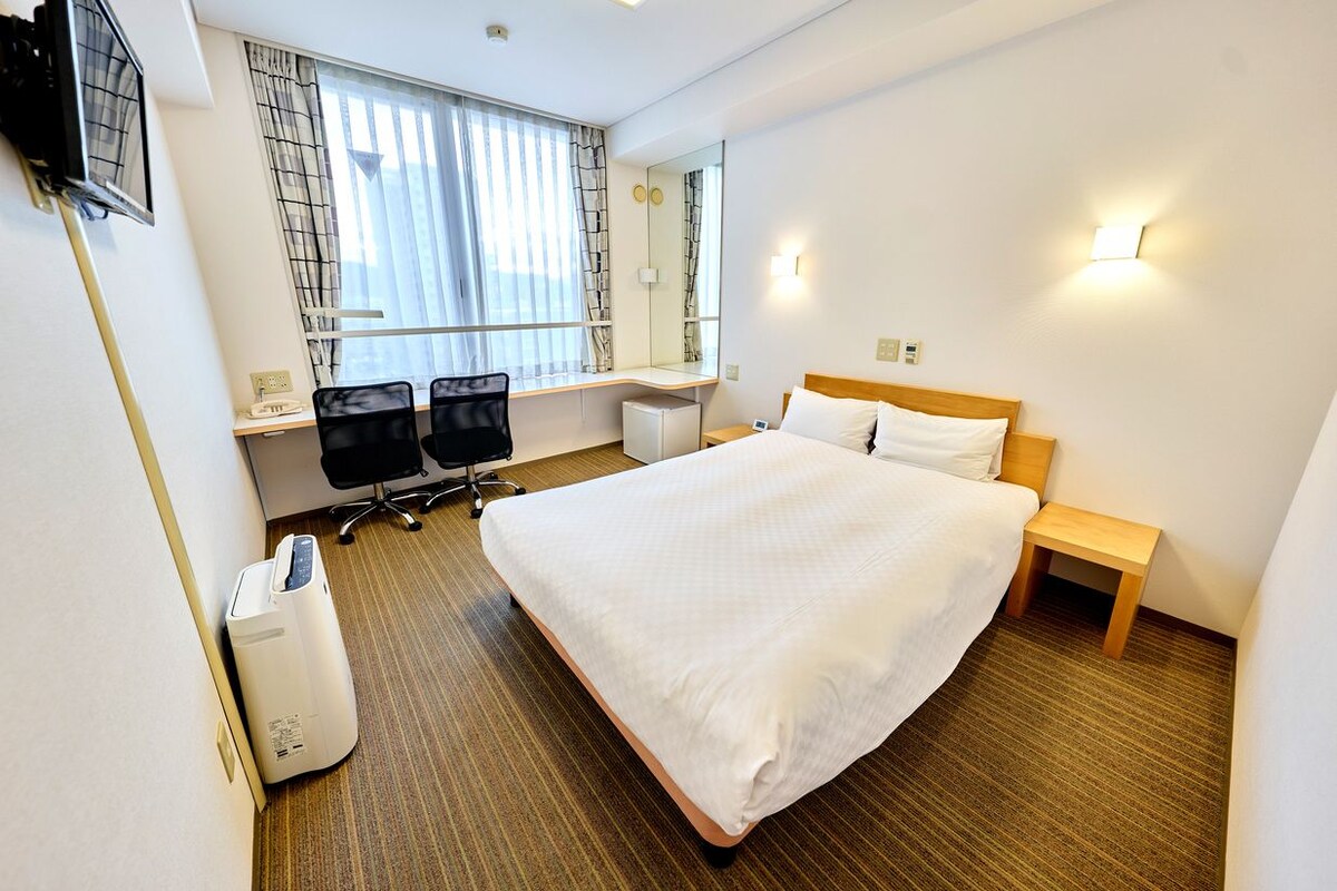 A hotel room with a large bed, a desk with a chair, a small bedside table, a trash bin, and a window with curtains.