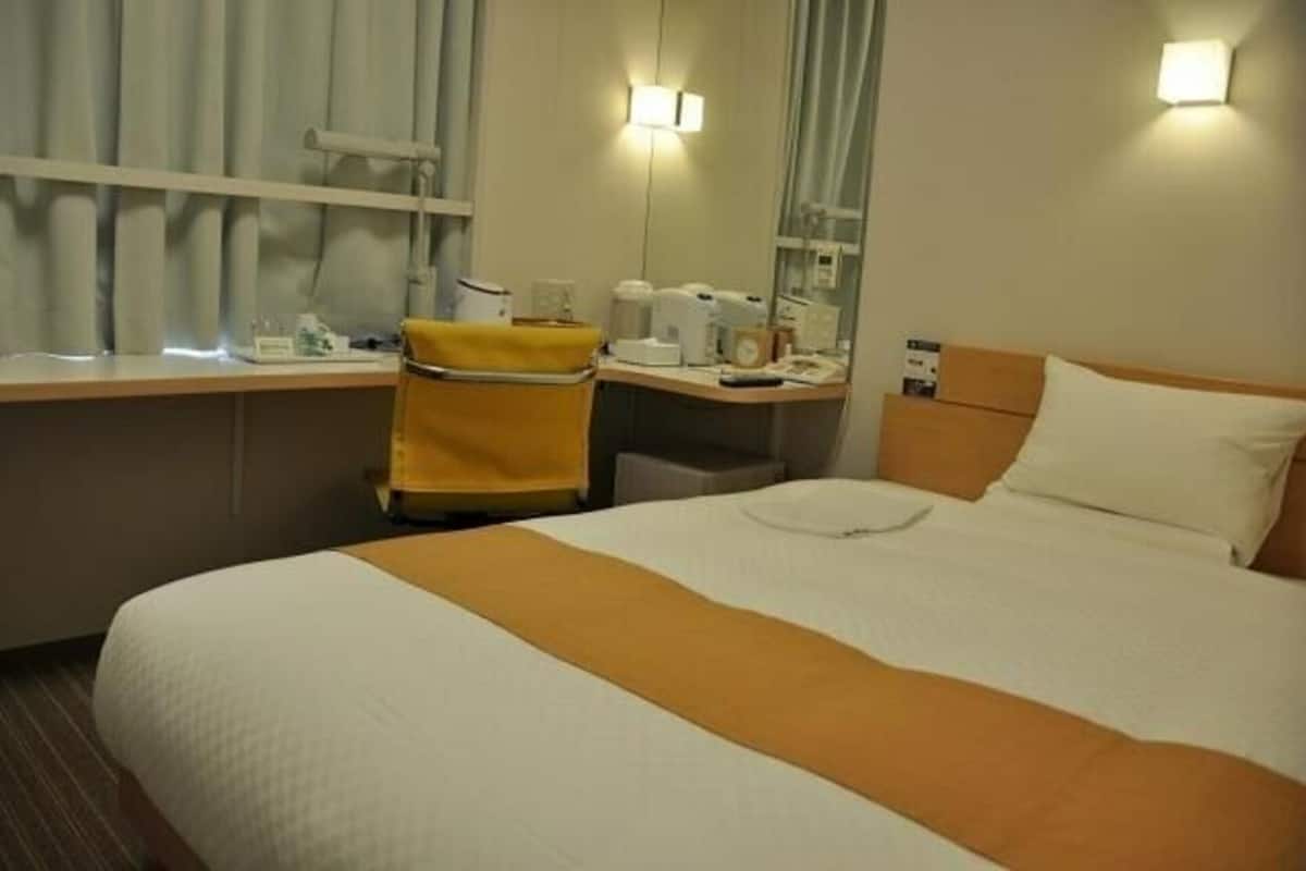 Une chambre d’hôtel avec un lit, un bureau, une chaise, un miroir et une fenêtre avec des rideaux.