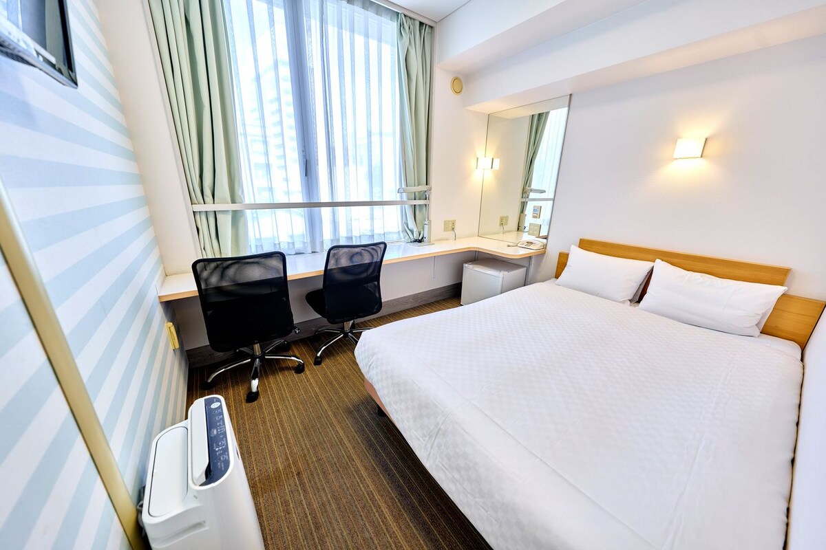 A hotel room with a large bed, a desk with a chair, a small refrigerator, a window with curtains, and a wall-mounted mirror.