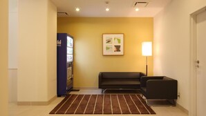 Een hotel lobby met een automaat, een bank, een lamp en een ingelijst kunstwerk aan de muur.