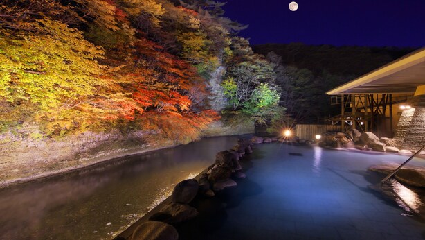 A nighttime view of a hot spring area with a river, rocks, and trees illuminated by colorful lights. - Yu No Mori Hotel Shidotaira (Hanamaki)