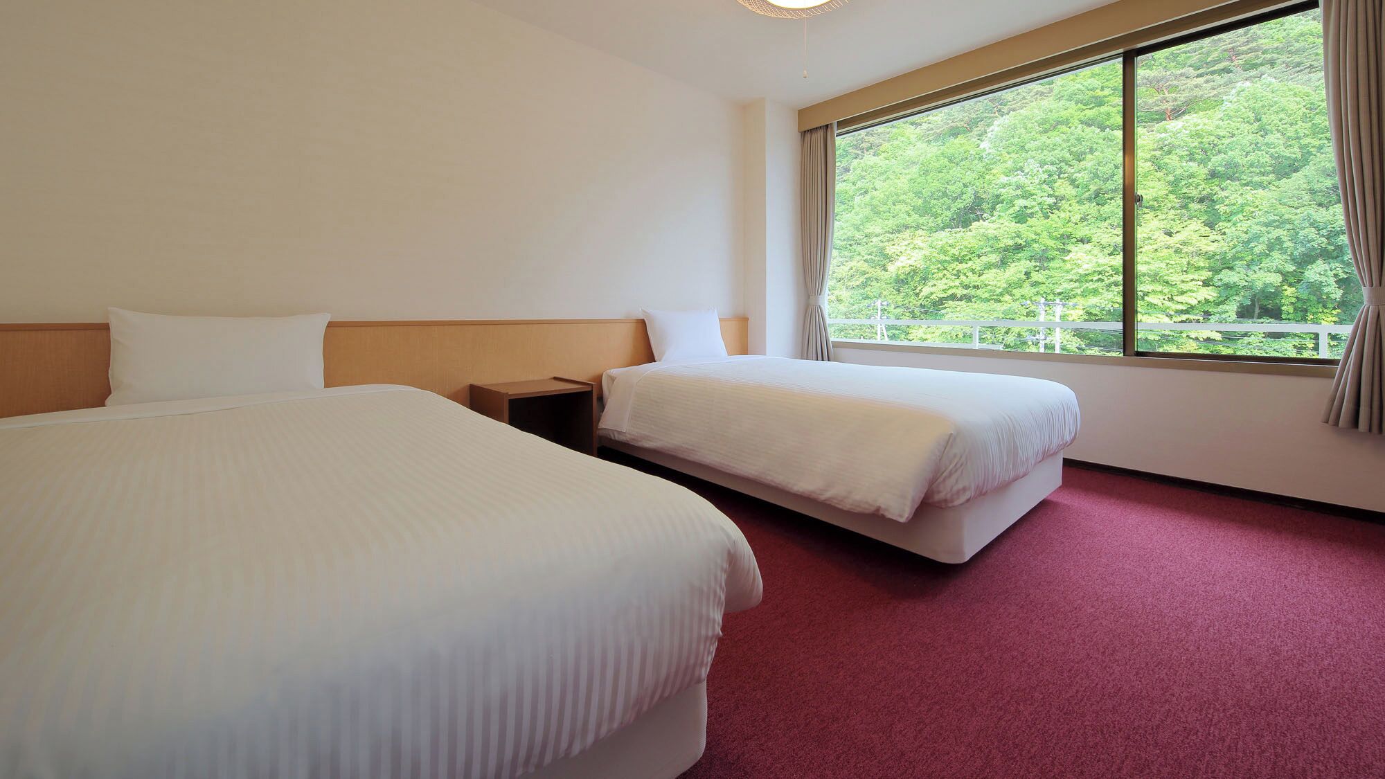 Een hotelkamer met twee bedden, een houten hoofdeinde, een nachtkastje, een groot raam met uitzicht op groen, en een rood tapijt.