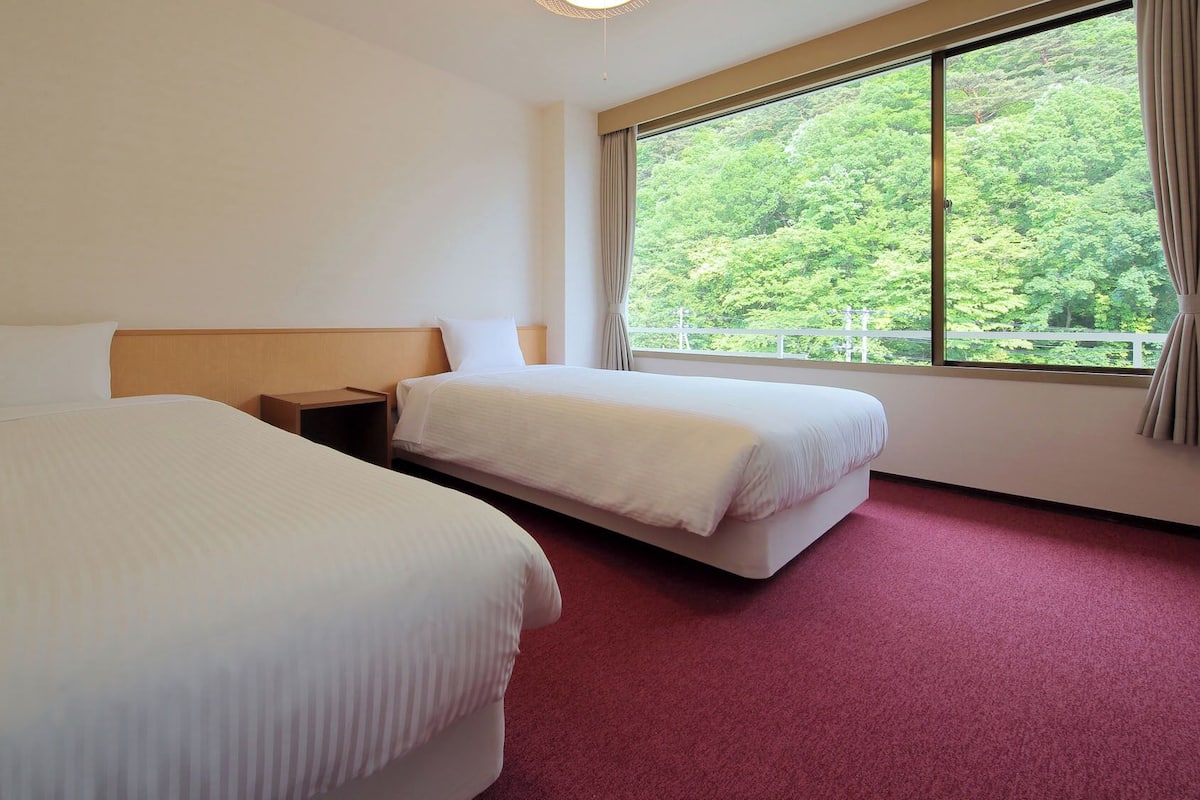 Ett hotellrum med två sängar, en sänggavel i trä, ett nattduksbord, ett stort fönster med utsikt över grönområden och en röd matta.