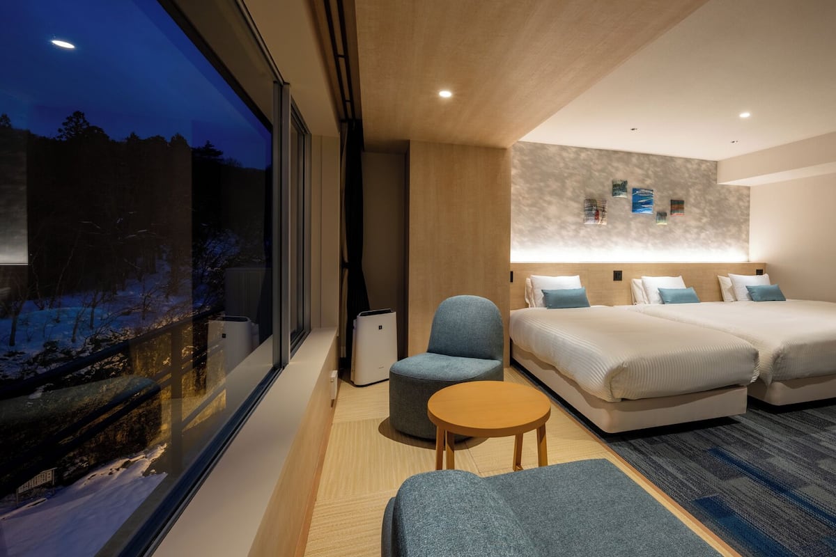 Ett modernt hotellrum med ett stort fönster, två sängar, ett litet bord och en stol.