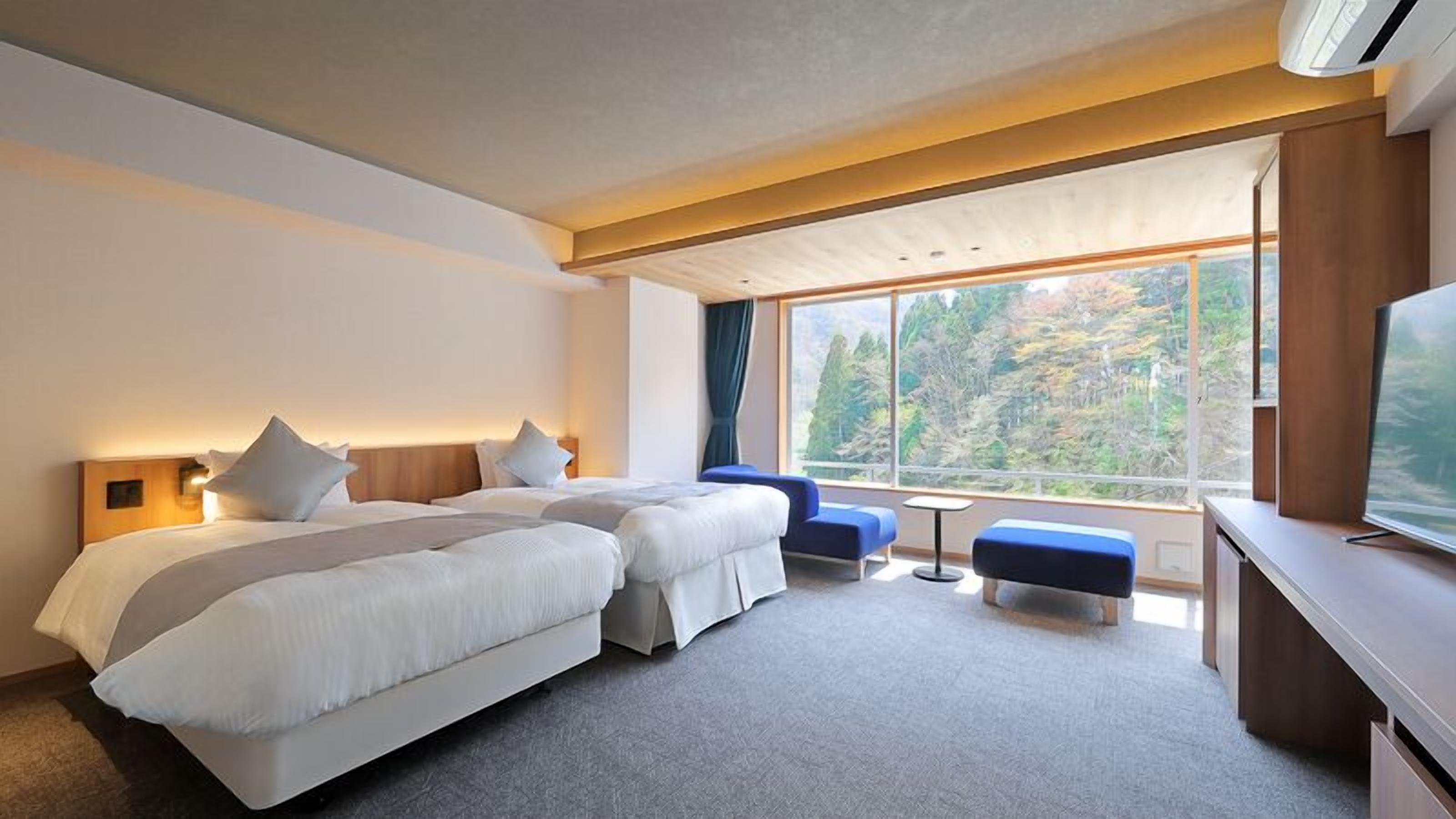 Hotelkamer met twee bedden, een bureau, een televisie en een groot raam met uitzicht op een bos.