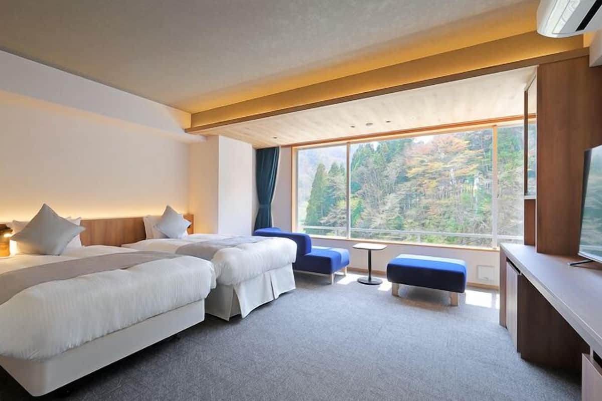 Ett hotellrum med två sängar, ett skrivbord, en TV och ett stort fönster med utsikt över skogen.