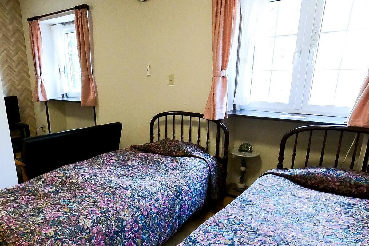 A room with two beds, a window with curtains, a mirror, and a small table with a clock.