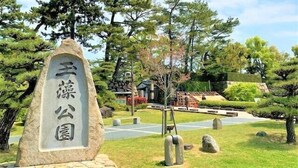 Un monument de pierre orné de caractères japonais, entouré d’un jardin bien entretenu où poussent différentes espèces d’arbres, avec un bâtiment traditionnel en arrière-plan.