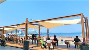 Un espace extérieur aménagé avec des tables et des chaises en bois, protégé par de grandes auvents, et offrant une vue sur une étendue d’eau où l’on aperçoit un navire de croisière au loin.