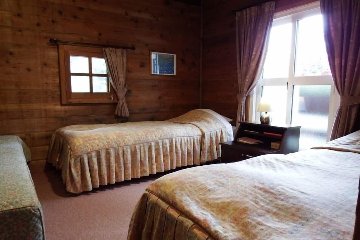 Quarto de cabana de madeira com duas camas, uma mesinha e uma janela com cortinas.
