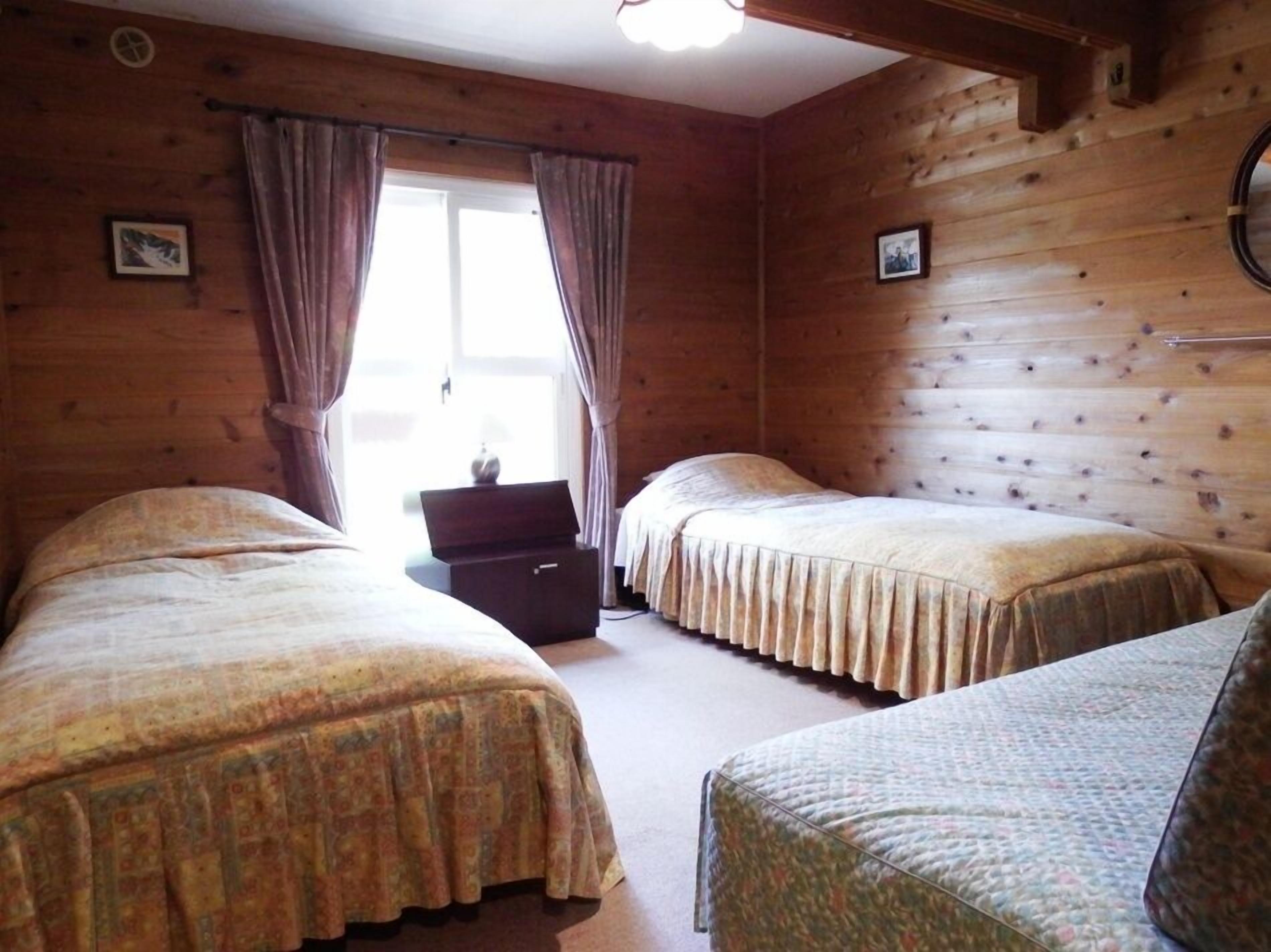 A room with two beds, wooden walls, a window with curtains, and a small table.