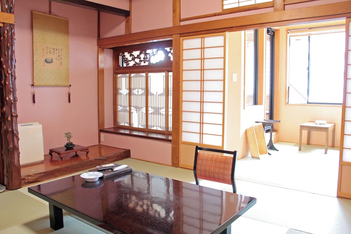 A traditional Japanese room with tatami flooring, a low table, a chair, sliding doors, and a view through a window.