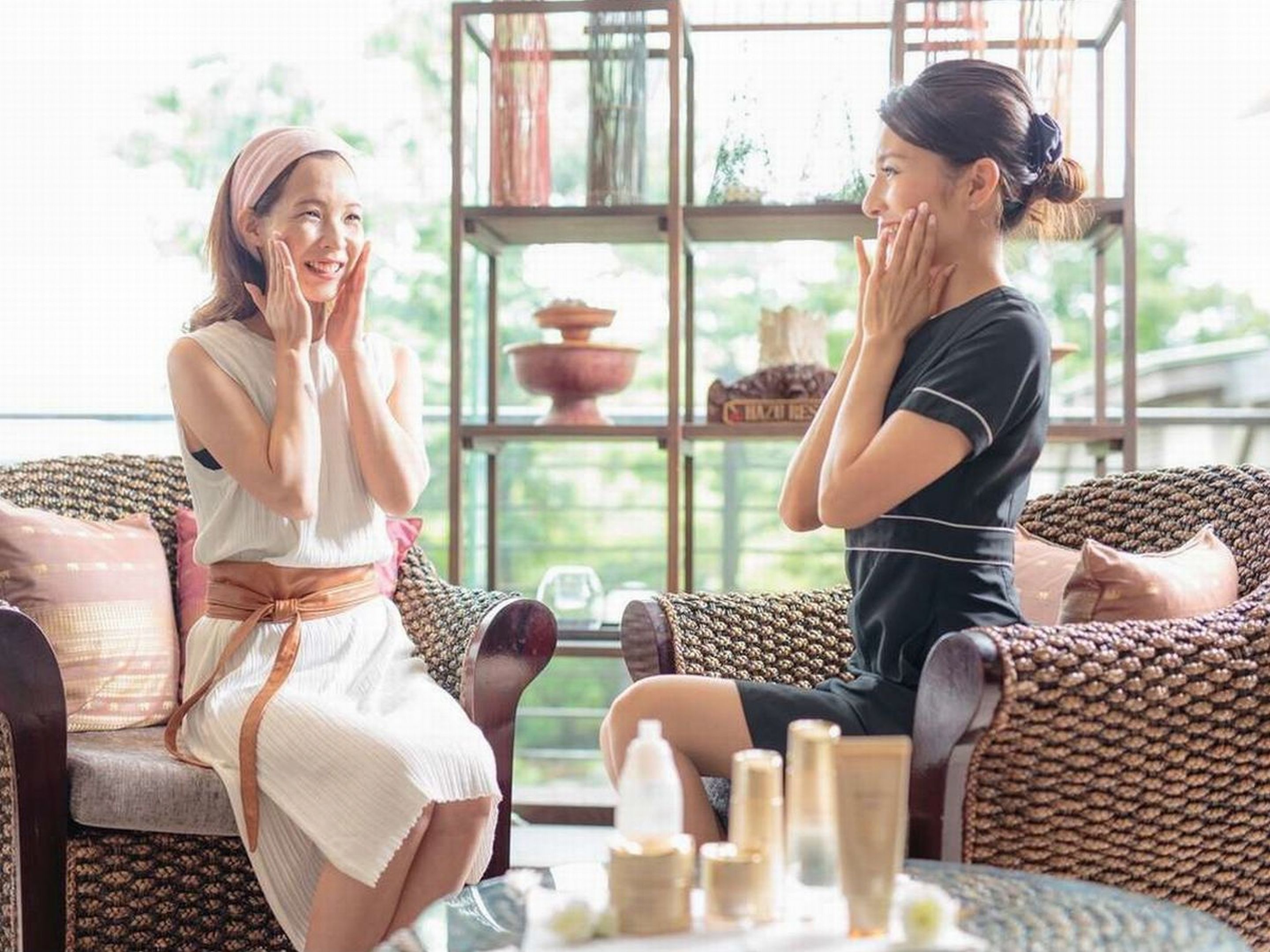 Two people sitting on wicker sofas in a well-lit room with a large window, a bookshelf, and a table with skincare products.
