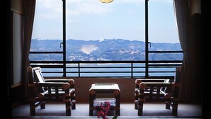 A room with a large window offering a view of a lake and mountains. There are two wooden chairs and a small table with a vase of flowers. - Wano Resort Hazu (Gamagori)