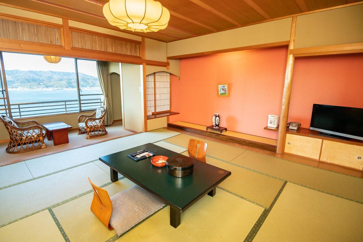 Ein traditioneller japanischer Raum mit Tatami-Böden, einem niedrigen Tisch mit Tee-Service, einem Fernseher und Blick aufs Meer.