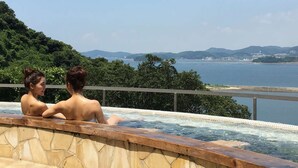 A hot tub with two people, surrounded by a stone wall and a view of a body of water and distant hills. - Wano Resort Hazu (Gamagori)