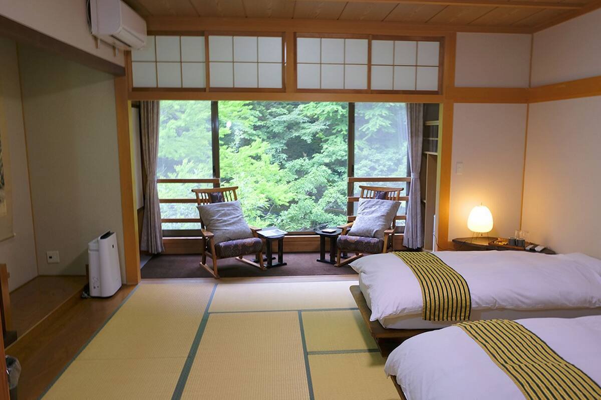 Een traditionele Japanse kamer met tatami vloerbedekking, een bed, twee schommelstoelen, een tafeltje en een raam met uitzicht op groen.