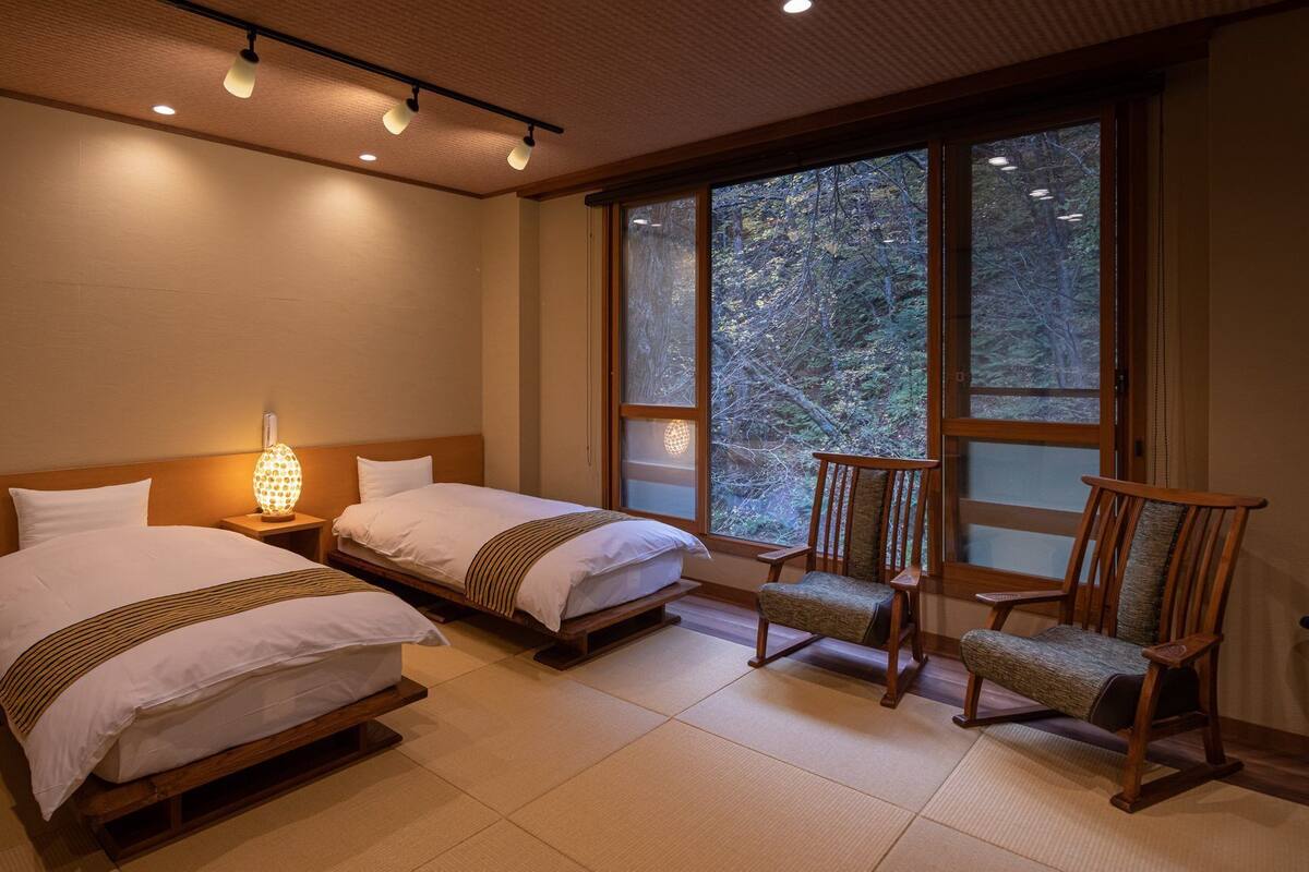 Hotelkamer met twee bedden, een nachtkastje met lamp, een houten stoel en een groot raam met uitzicht op bomen.