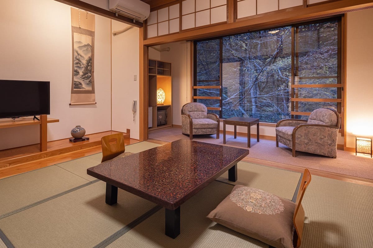 Een traditionele Japanse kamer met tatami vloerbedekking, een lage tafel, stoelen, een televisie en een groot raam met uitzicht op bomen.