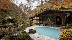 A rustic cabin with a stone wall, a wooden deck, and a natural pool surrounded by autumn foliage. - Tsuruya Ryokan (Matsumoto)