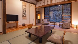 A traditional Japanese-style room with tatami flooring, a low table, chairs, a television, and a large window with a view of trees. - Tsuruya Ryokan (Matsumoto)