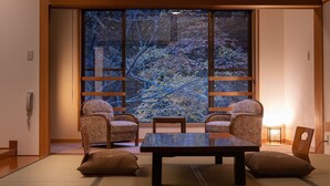 A traditional Japanese room with tatami flooring, a low table, two chairs, and a large window with a view of trees. - Tsuruya Ryokan (Matsumoto)